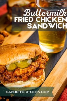 a fried chicken sandwich with pickles on a cutting board next to a glass of beer