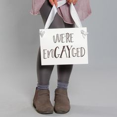 a woman holding a sign that says we're engaged in front of her legs