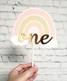 a hand holding a cake topper with the word one on it and a rainbow