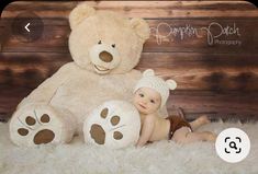 a teddy bear sitting next to a baby doll on a fluffy white rug in front of a wooden wall