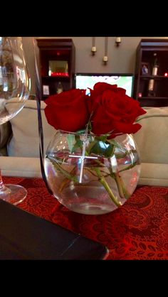 two red roses in a clear vase on a table with wine glasses and a book