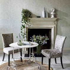 two chairs and a table in front of a fire place with flowers on the mantle