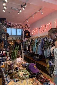 Girl browsing in thrift shop with neon sign  lots of accessories on the table and racks of clothes against back wall #charityshop #thriftshop #fashion #slowfashion #clothing #diyclothing #outfitinspiration Thrift Store Aesthetic Pics, Small Town Clothing Store, Aesthetic Thrifted Outfits, Cute Thrift Shop, Clothing Store Decor Ideas, Thrifting Aesthetic Pics, Cool Boutique Interiors, Vintage Store Decor