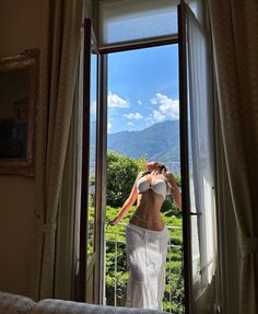 a woman standing in front of an open window looking out at the mountains and greenery