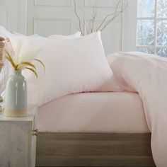 a bed with pink sheets and pillows next to a vase filled with flowers in front of a window