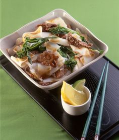 a dish with noodles, meat and vegetables on a black tray next to chopsticks