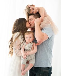 a man and woman holding a baby in front of a white background with the child's mother kissing her cheek