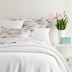 a white bed topped with lots of pillows next to a vase filled with pink flowers