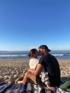 a man and woman sitting on top of a blanket at the beach kissing each other