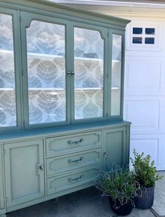 an old china cabinet painted green with white lace on the glass doors and drawers, next to a potted plant