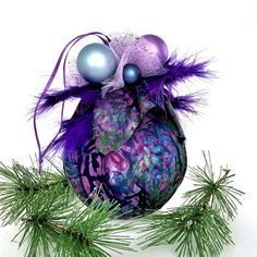 a christmas ornament with purple feathers and balls on top of it, surrounded by pine branches