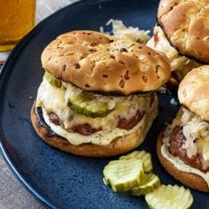 three hamburgers with pickles and cheese on a blue plate next to a glass of beer