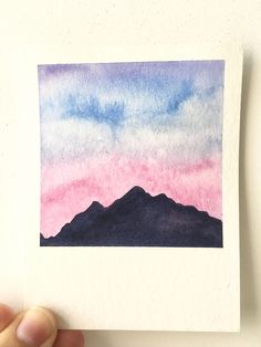 a hand holding up a piece of paper that has watercolor on it with mountains in the background