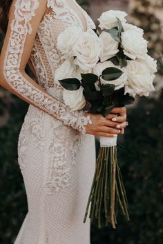 a woman in a wedding dress holding a bouquet of white roses and greenery with her hands on her hips