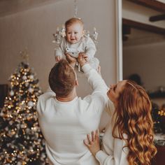 a man holding a baby up to his face while standing next to a woman in front of a christmas tree