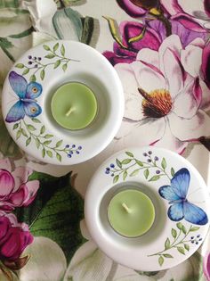 two tea lights sitting on top of a floral table cloth with butterflies painted on them