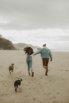 a man and woman running on the beach with two dogs behind them, holding hands