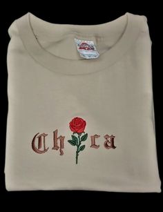 "Embroidered Chica T Shirt w/Red Rose Short Sleeve, Crew Neck, beige in color. The embroidered Chica is 5\" in length the rose is 2.75\" tall. (Adjusted to the shirt size) ] 100% cotton  Set-in rib collar with shoulder-to-shoulder taping Double-needle sleeve and bottom hem Preshrunk to minimize shrinkage. Wash: Cold water, gentle cycle/delicate Dry: Low heat or drip dry Iron: Cotton setting \"Colors May Vary\"" Red Embroidered Graphic Tee, Casual Red T-shirt With Embroidered Graphics, Casual Red Embroidered T-shirt, Red Embroidered Crew Neck T-shirt, Pink Crew Neck T-shirt With Custom Embroidery, Zoot Suit, Brown Pride, Latest T Shirt, Pride Tshirts