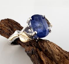 This beautiful Hand Cut Blue Sapphire Engagement Ring with two tone gold leaves, a very unique One-of-a-kind Engagement Ring. Description: -Hand Cut 9.71mm x 6.43mm Hand Cut Blue Sapphire  -The Band is cast in 14k White Gold with textured bark markings with 14k Yellow Gold Leaf accents. 9.71mm x 6.43mm Hand Cut Blue Sapphire Overall Ring Width: 9.44mm Bottom Band Width: 2.27mm Our Refined Gold and Gemstones are all Cruelty-Free. Additional Raw Hand Cut Gemstone Wedding Rings: https://www.etsy.co Gold Engagement Ring Sapphire, Sapphire Gold Engagement Ring, Branch Wedding Band, Sapphire Flower Ring, Twig Wedding Band, Teal Sapphire Ring, Engagement Ring Sapphire, Blue Sapphire Engagement Ring, Gemstone Wedding Rings