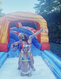 two women in swimsuits are sitting on an inflatable slide while another woman holds her arm out