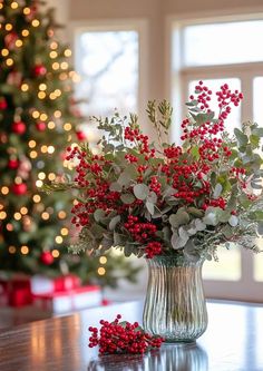 a vase filled with red berries and greenery on top of a table next to a christmas tree