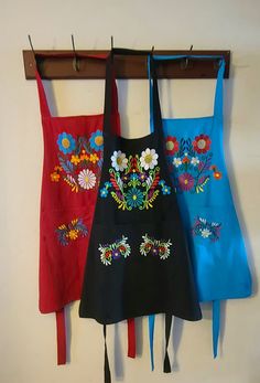 three aprons hanging on a wall with tassels attached to the hooks and two are decorated with flowers