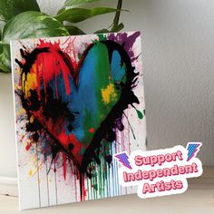 a colorful heart painted on top of a white canvas next to a potted plant