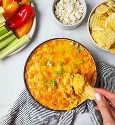 a person holding a tortilla chip in front of a bowl of dip and vegetables