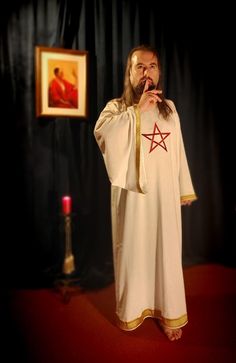 a man with long hair standing in front of a painting and holding his hands to his mouth