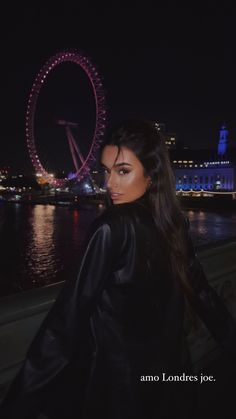 a woman standing next to the water with a ferris wheel in the background at night