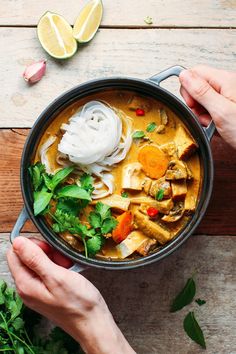 two hands holding a bowl of soup with noodles and vegetables on the table next to sliced lemons