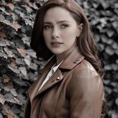 a woman wearing a brown leather jacket standing in front of a wall covered with leaves