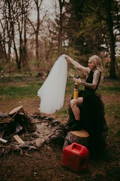 a woman in a black dress sitting next to a fire pit holding a white wedding dress