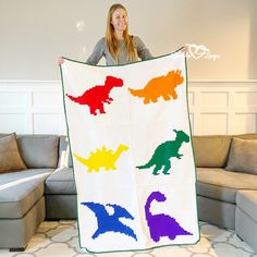 a woman holding up a colorful dinosaur crochet quilt in front of a couch