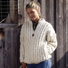 a woman standing in front of a wooden fence wearing a white sweater and jeans with her hands on her hips