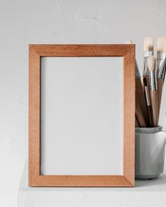 an empty wooden frame sitting on top of a table next to paintbrushes and a cup