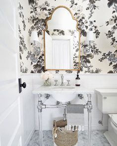 a white bathroom with floral wallpaper and mirror above the sink, along with a wicker basket on the floor