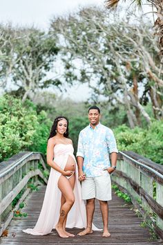 a pregnant couple standing on a wooden bridge in front of trees and bushes, posing for the camera