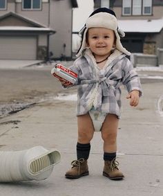 a baby in a hat and diaper standing on the sidewalk next to a trash can