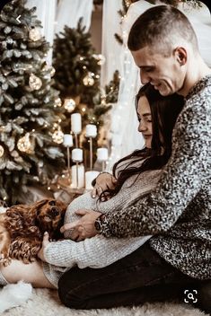 a man and woman sitting on the floor with their dog in front of a christmas tree