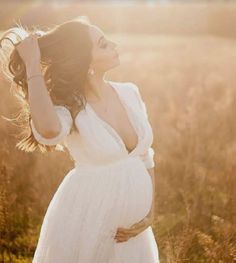 a pregnant woman in a white dress standing in a field with her hands on her head