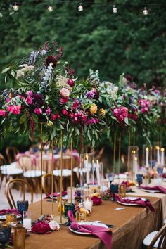 a long table topped with lots of purple and red flowers next to tall gold candles