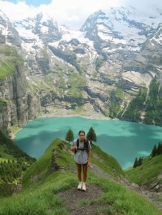 a woman standing on top of a lush green hillside next to a lake with snow covered mountains in the background