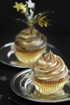 two cupcakes with gold frosting sitting on top of silver plated plates