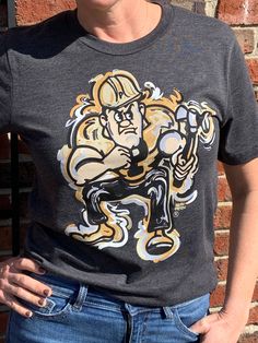 "Justin Patten brings his unique, artistic style to Purdue Pete with this exclusive Purdue University licensed apparel. His artwork is printed on a high-end Bella + Canvas tee with a water-based 6 color print, to produce a soft, cozy tee with a vibrant image. The water-based screen printed image is like no other. You will love the feel of this shirt from day one, and it will look and feel great after years of wear. Sizing: The unisex tees are a traditional cut shirt for men and women. They run t Graphic Tee Tops With Custom Artwork For Fans, Custom Artwork Crew Neck Top For Fan Merchandise, Crew Neck Tops With Custom Artwork For Fan Merchandise, Fire Hat, West Lafayette, Purdue University, Cut Shirt, Artistic Style, Bella Canvas Tees