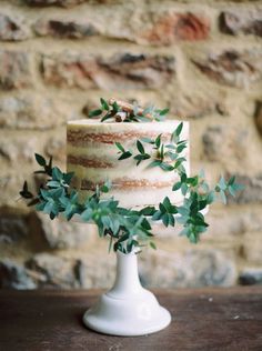 a three tiered wedding cake with greenery on the top and bottom, sitting in front of a brick wall