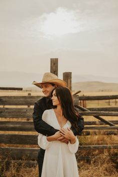 a man and woman standing next to each other in front of a wooden fence on a farm