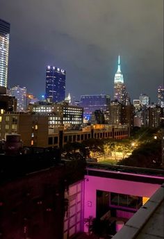 the city skyline is lit up in pink and blue lights at night, as seen from an apartment balcony