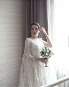 a woman in a white wedding dress holding a bouquet and standing next to a window