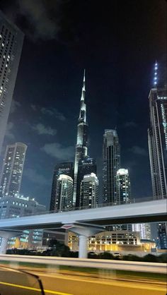 the city skyline is lit up at night with skyscrapers in the foreground and cars passing by